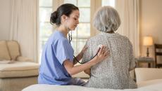 Two people sitting on a bed in a home with one being a healthcare provider checking the other one's vitals