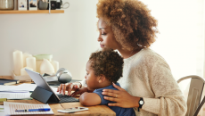 A woman holds a baby while using a tablet