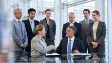 Several business people standing around a table watching two people sitting down shaking hands