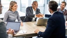 Five businesspersons sitting around a table with two of them shaking hands