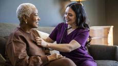 Healthcare provider sitting on a couch with a patient in their home checking their vitals. 