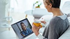 Person sitting on a couch holding a coffee cup while talking to a telehealth provider on their computer