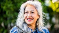 Person with long gray hair smiling while standing in front of trees