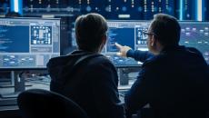 Two people sitting in front of three computer monitors analyzing data on the screen