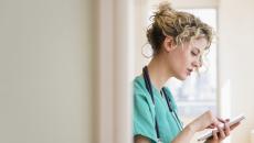 Healthcare provider standing in a hallway wearing green scrubs and a stethoscope around their neck while looking at a tablet