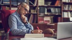 Person sitting at a desk writing something down on a piece of paper with a bookshelf behind them