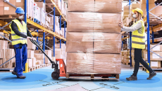 Two workers in vests and masks move a pallet of boxes with a machine lift.