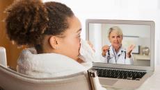 Person sitting in a chair while blowing their nose and talking to a healthcare provider on a computer