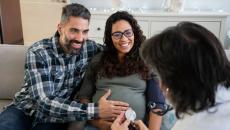 Two people sitting on a couch with one rubbing the other's stomach while both speak to a healthcare provider