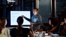 Person presenting in front of other people while standing next to a screen in a dark room