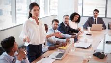 Numerous people in a board room sitting around a table with one person standing up