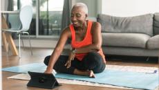 A woman sitting on a yoga mat using a tablet before she exercises