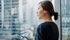 A woman holding a smartphone looking out a window.