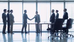 Two people shaking hands in front of a big window with six other people watching