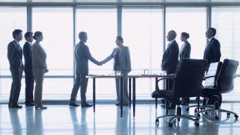 Two people shaking hands by a window while other people watch