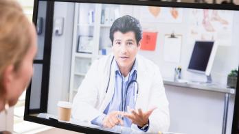 Healthcare provider on a computer screen talking to someone sitting at a desk