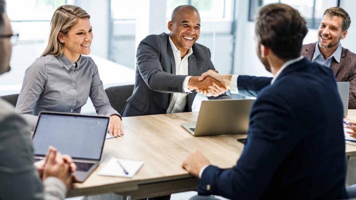 Five businesspersons sitting around a table with two of them shaking hands